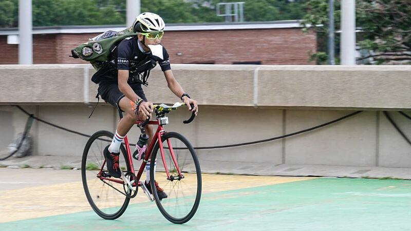 Seorang kurir sepeda mengenakan helm dan kacamata tengah mengayuh sepeda sambil membawa tas dan secarik kertas